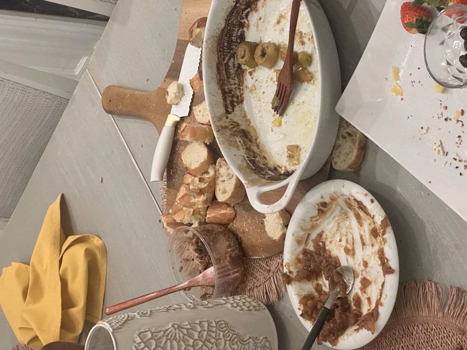 overhead of demolished baked brie in oval dish w/ olives + jam + bread cubes on cutting…