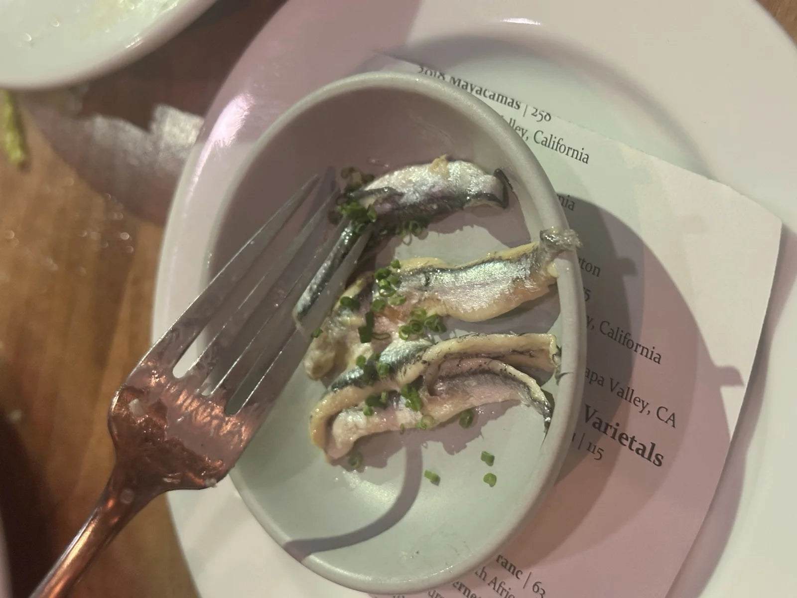 small plate of white anchovies / boquerones w/ herbs, being lifted w/ a copper-handle f…