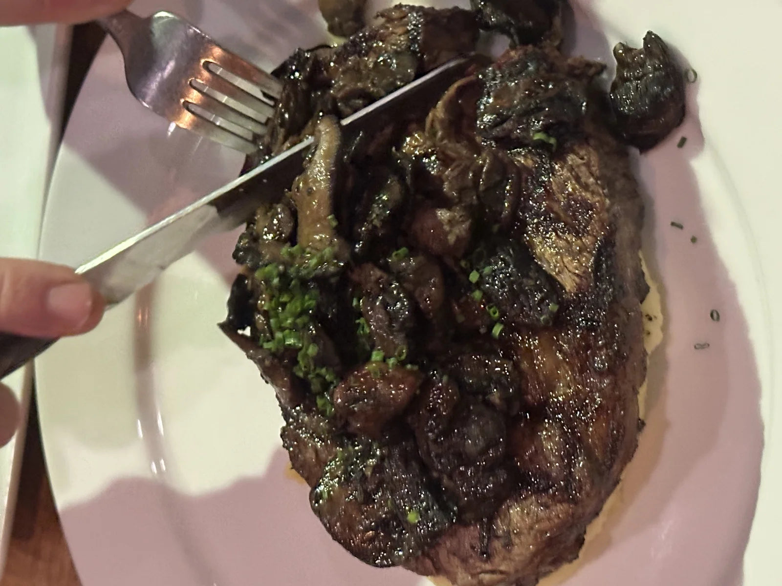 hero shot of a ribeye topped w/ pan-roasted mushrooms + chives being cut into on a pink…