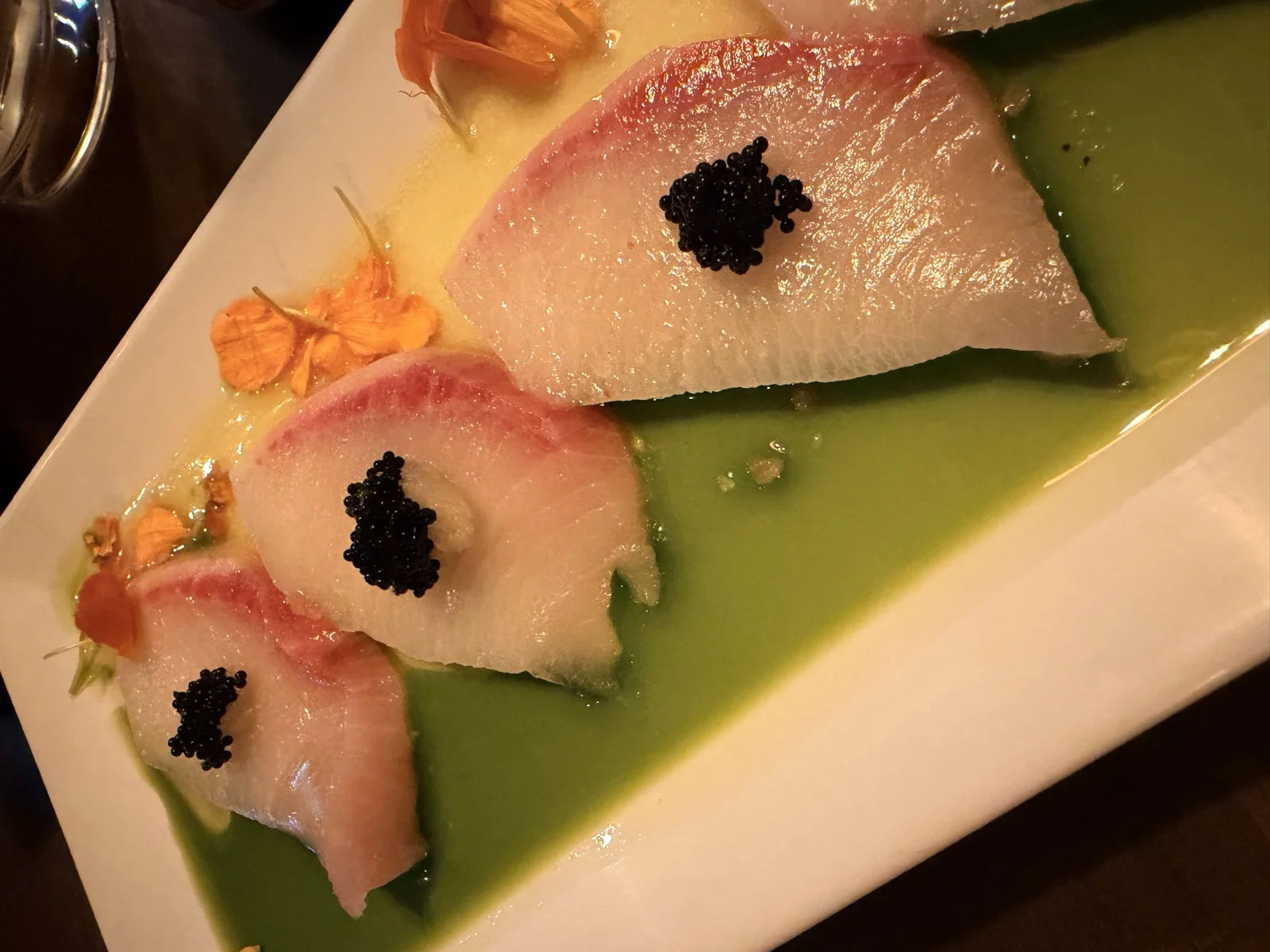 three slices of hamachi crudo w/ caviar dots on a green olive-oil/tomatillo pool at a r…