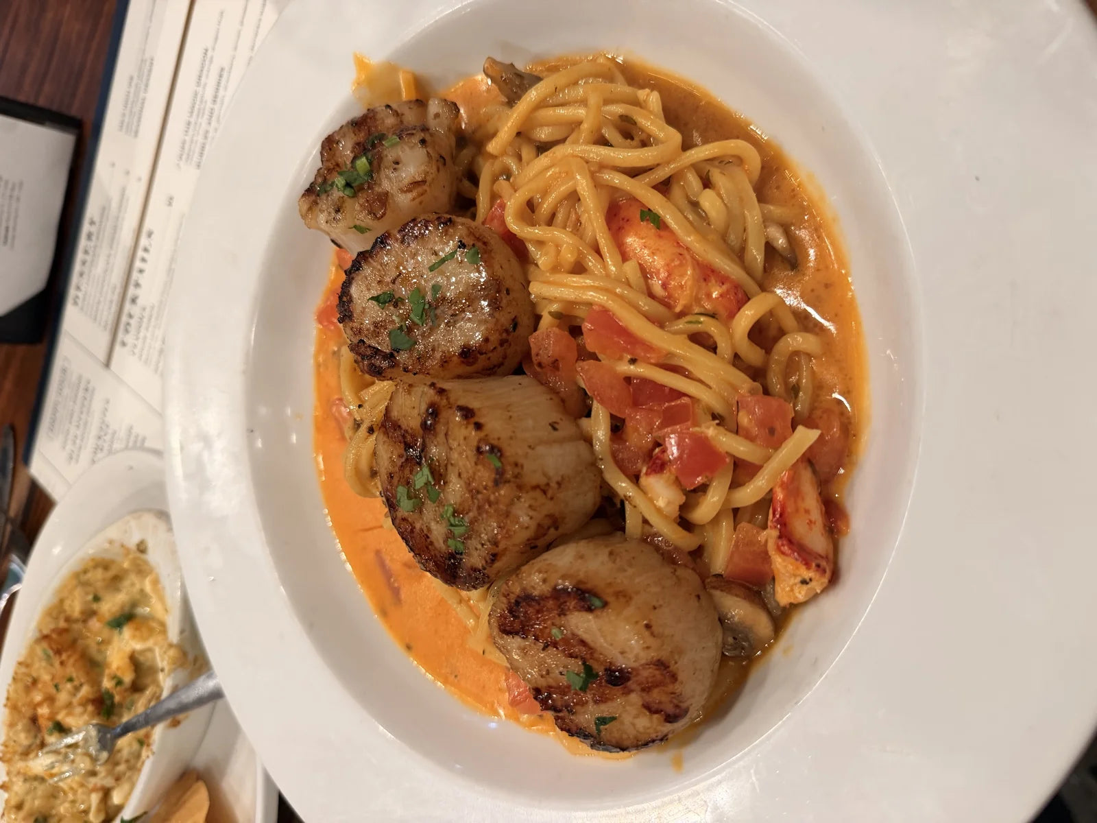 seared scallops over lobster + tomato cream linguine on a white plate, side of crab-top…